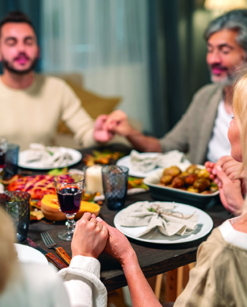 Contemporary family of four sitting by served festive table in living-room, holding by hands and saying thanks to Lord while keeping eyes closed