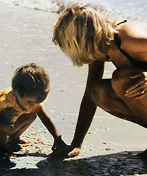 A little boy plays in sand while his mother kneels over him and helps.