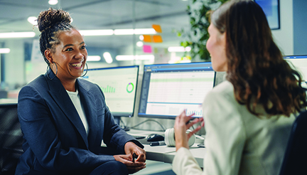 Two Female Colleagues Fondly Talk to Each Other, Laugh and Smile while Working on Computers in Diverse Modern Business Office. Experienced Manager and Young Employee Discuss a Fun Analytical Project.