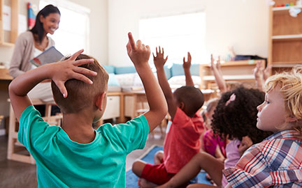 A teacher reading to elementary aged children sitting on the floor, raising their hands